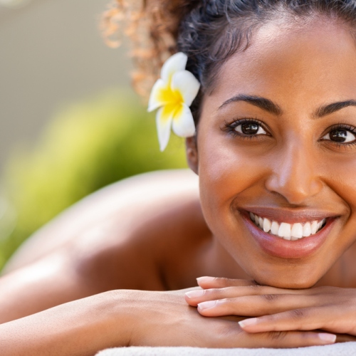 Closeup face of young african american woman in a wellness center ready for massage therapy. Portrait of beautiful girl lying down on massage table at spa resort. Smiling girl looking at camera while awaiting beauty treatment.