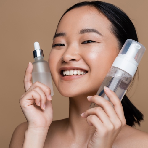 Positive asian woman with naked shoulders holding serum and face cleanser isolated on brown